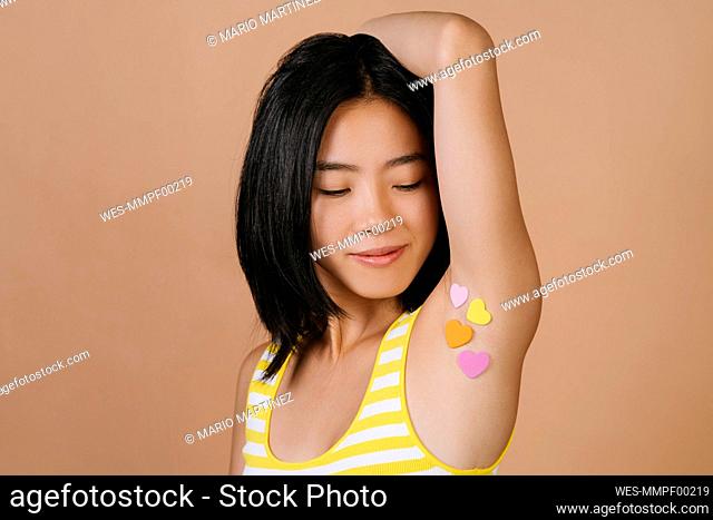 Young woman with heart shape stickers on armpit against brown background