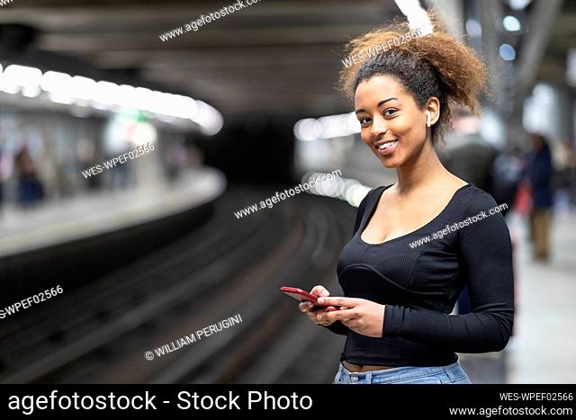 Portrait of happy young woman with cell phone and earbuds at subway station platform