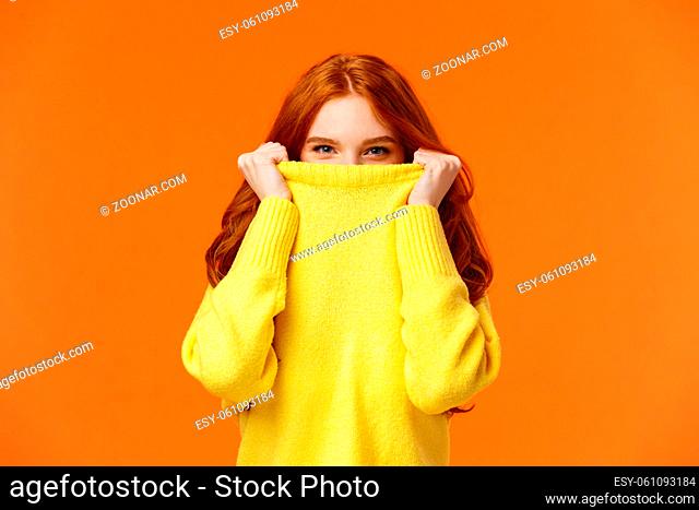 Girl hiding from winter cold in her warm soft sweater. Cute redhead woman pulling collar on nose and peeking at camera with smiling eyes, glancing at you happy