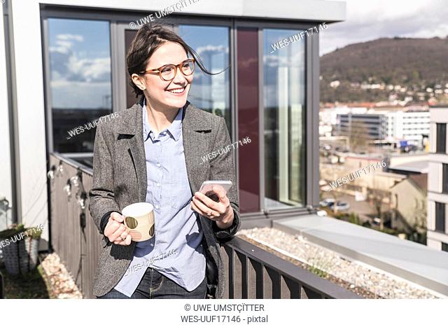 Smiling businesswoman with cell phone and cup of coffee on roof terrace