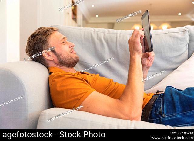 Caucasian man lying on couch, smiling and using tablet at home