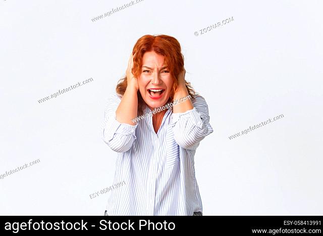 Portrait of distressed and upset redhead female in shirt, screaming in panic, cover ears with hands concerned, standing anxious and insecure over white...