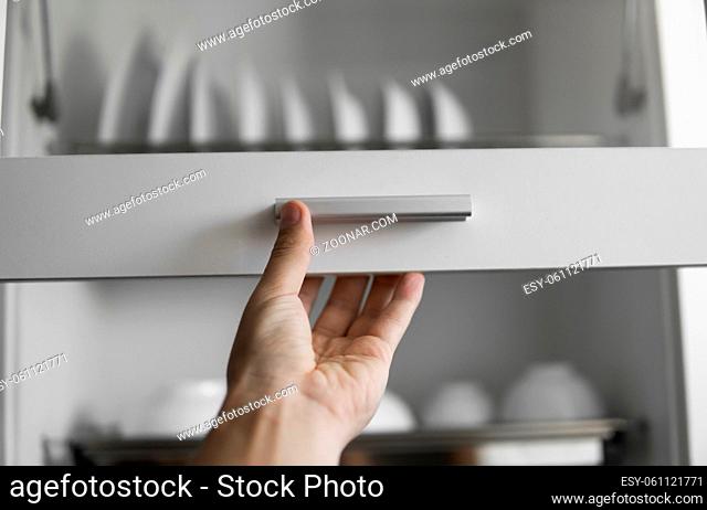 Hand holds the door of the dryer with utensils. Dish drying metal rack with white clean plates. Traditional comfortable kitchen