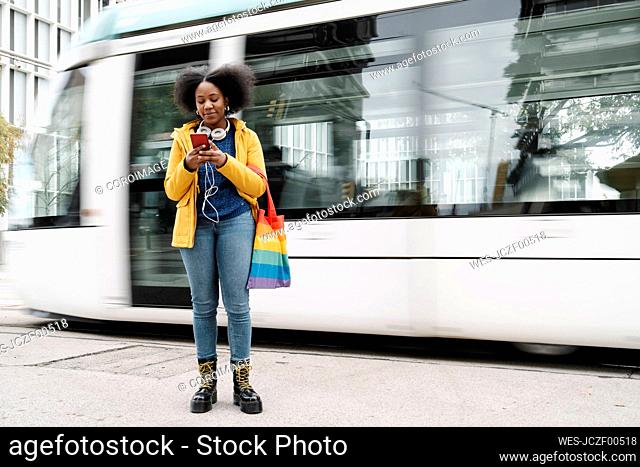 Young woman text messaging on mobile phone while standing at footpath against tram