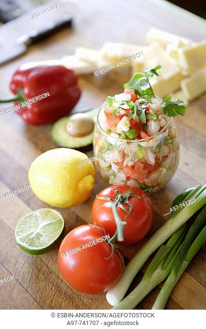 Glass of ceviche, tomatoes, lemon, scallions & lime in foreground, sliced cheese, bell pepper, & avocado in background
