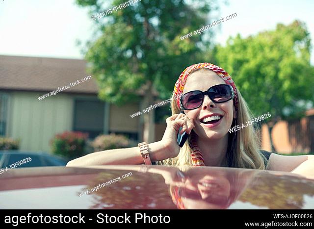 Laughing woman talking on mobile phone leaning on car during sunny day