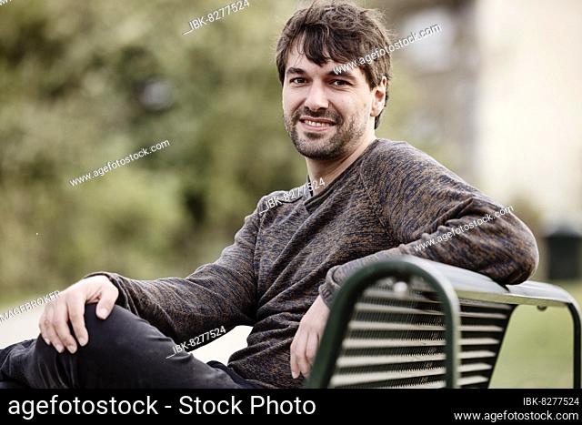 Junger Mann draußen auf einer Parkbank sitzend, Portrait, Köln, Nordrhein-Westfalen, Deutschland
