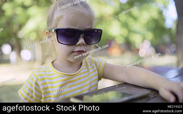 Little girl in mom's sunglasses is learning foreign language by repeating words from mobile phone. Close-up portrait of child girl sitting in city park and...