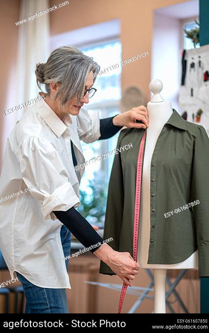 Measurements. Gray-haired woman standing sideways to camera touching shirt with measure on dummy in sewing workshop during day