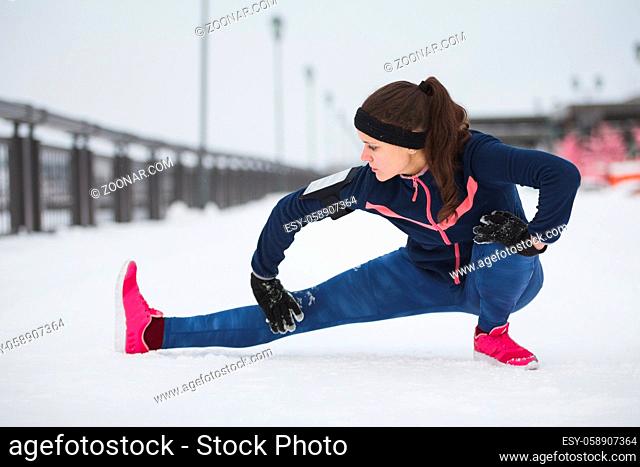 Woman runner stretching legs before run at snow winter promenade. Fitness concept., telephoto