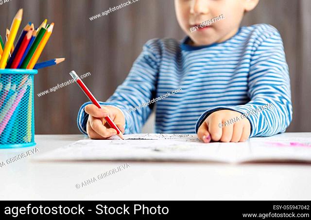 Little child at home drawing on the paper. Boy sitting on the chair at table. Five years old kid writing at desk in room with wooden background