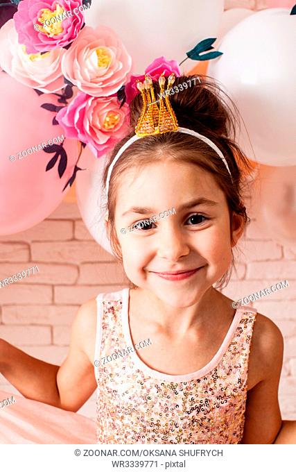 The beautiful little girl with crown over pink girly balloon and paper flower decorations. Little princess is celebrating