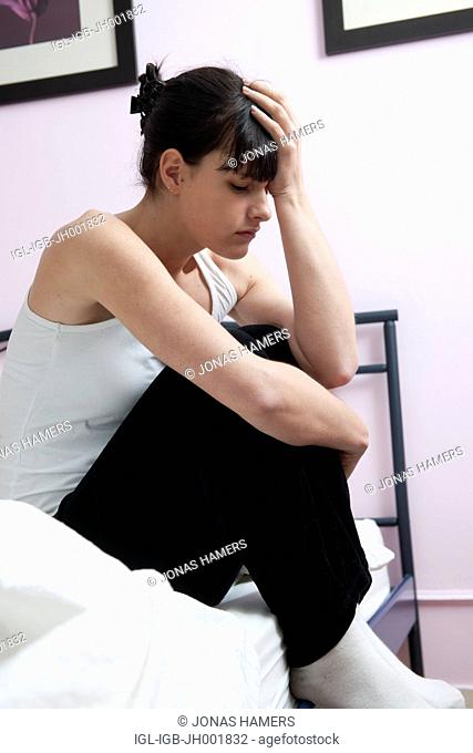 This picture shows a young caucasian woman with brown hair as she sits on her bed feeling ill or sick, tired or anxious