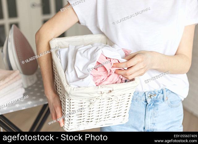 Wicker rustic basket with washed clothes in woman's hand ready to ironing. Close up view on basket in hands. Woman in living room