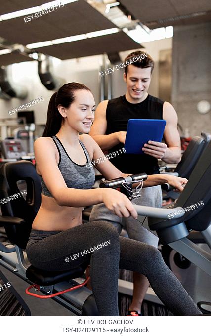sport, fitness, lifestyle, technology and people concept - happy woman and trainer with tablet pc computer working out on exercise bike in gym