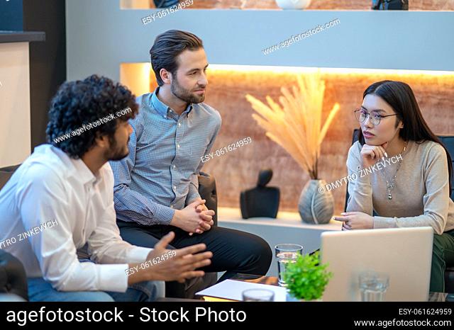 Two male office employees seated on the sofa staring at their pensive young female colleague