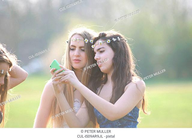 Two teenage girls wearing daisy chain headdresses reading smartphone texts in park