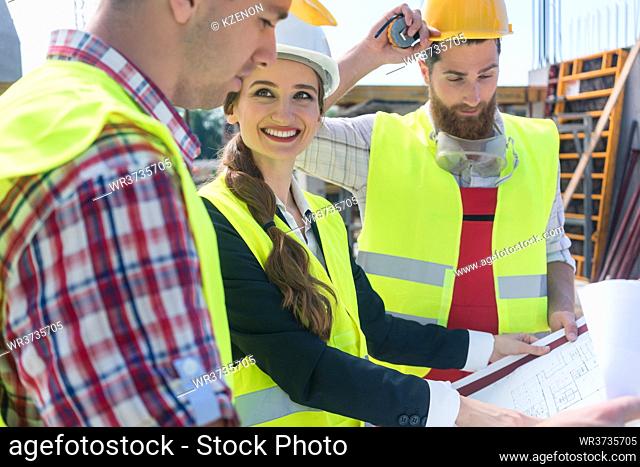 Young and confident female architect smiling while sharing ideas about the plan of a building under construction outdoors with two blue-collar workers
