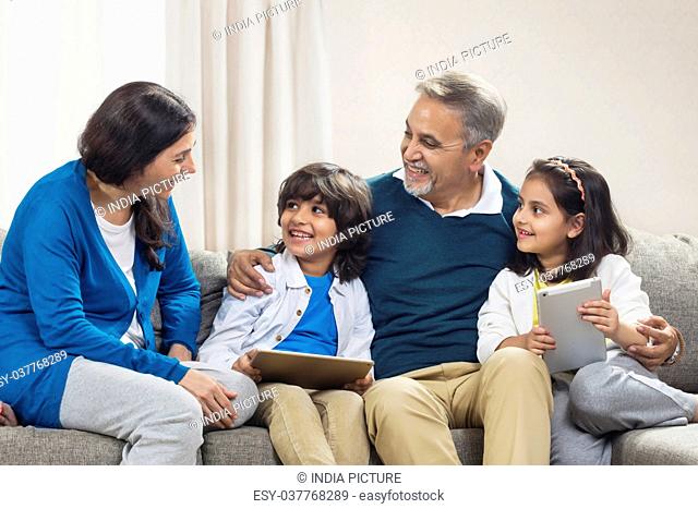 Smiling grandchildren using digital tablet with grandparents sitting on sofa