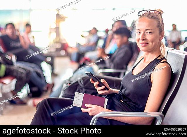Casual tanned blond female traveler smilling while holding cell phone, passport and boarding pass, waiting to board a plane at the departure gates at the asian...
