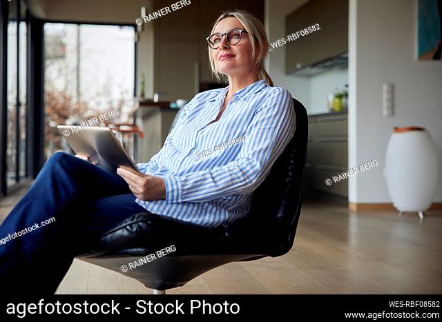 Thoughtful woman wearing eyeglasses sitting with tablet PC on chair at home