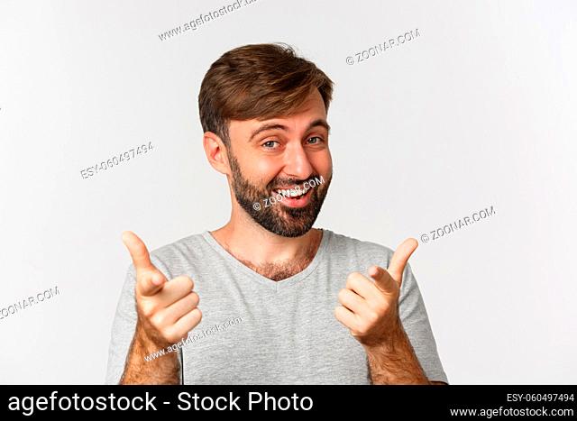 Close-up of handsome adult man with beard, smiling and pointing fingers at camera, standing in gray t-shirt over white background