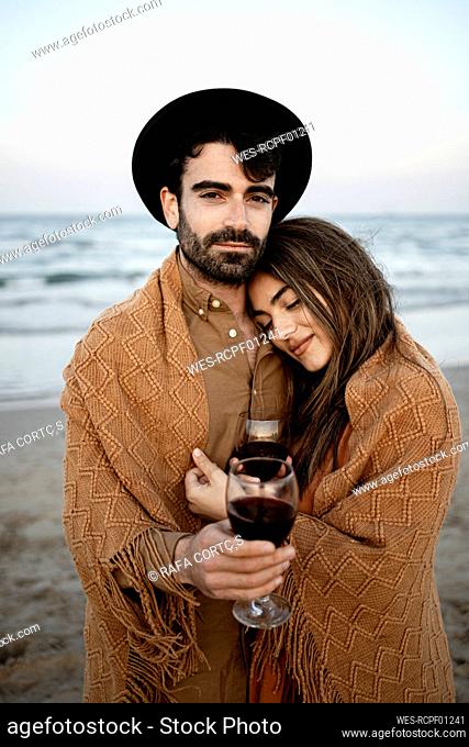 Boyfriend wrapped in blanket with girlfriend holding wine at beach