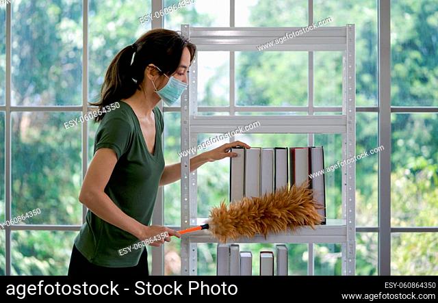 Asian housewife wearing face mask while cleaning the book shelf in living room with a feather duster