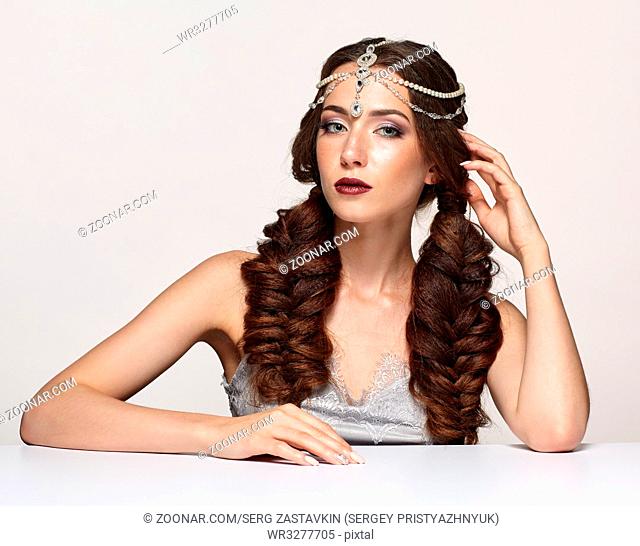 Beauty portrait of young woman in pearl diadem. Brunette girl with braids hairdo and day female makeup sit at white table on gray background