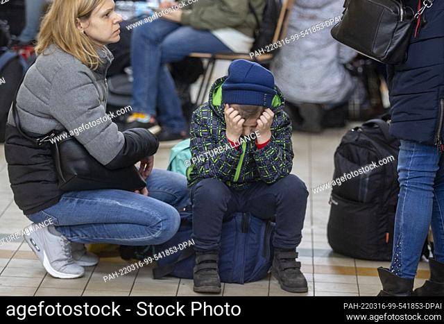 16 March 2022, Poland, Przemysl: A little boy in a cap sits exhausted on a travel bag in the train station in Przemysl, his mother squats next to him