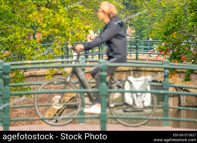 Netherlands. Summer day in Amsterdam. Defocused female cyclist on a small bridge over a canal against the backdrop of lush green park foliage