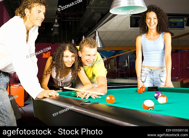 Small group of teenagers standing at pool table. Two of them are playing bliiard