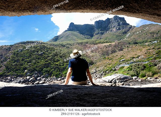 Man wearing a hat sits on a rock looking out at granite mountains, Llandudno cove beach, Atlantic Ocean, between Camp's Bay and Hout Bay, Cape Town