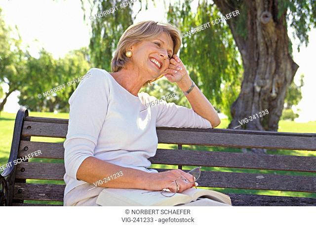 portrait, park-scene, blond woman, middle of 50, wearing bright trousers and top, sits relaxed with a book and her glasses in her hand on a bench under a tree...