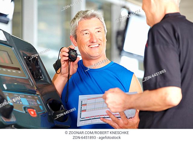 Fitnesstrainer steht mit Stoppuhr bei Senior am Laufband im Fitnesscenter