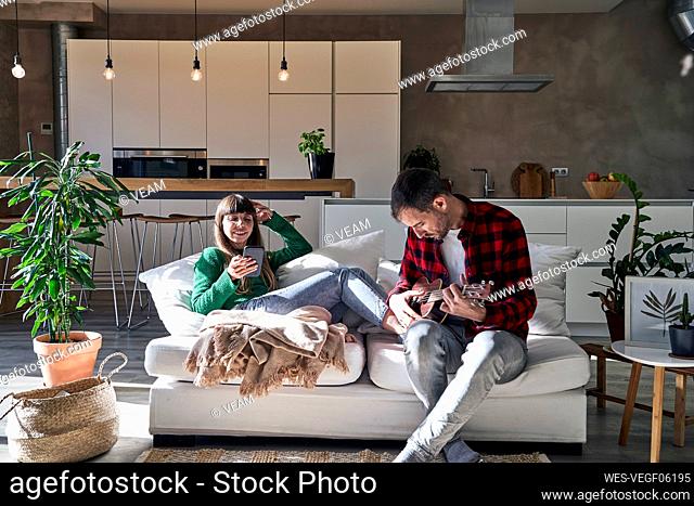 Man playing ukulele sitting by woman using smart phone on couch at home