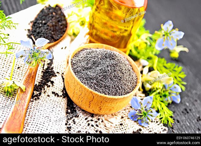 Flour of black caraway in a bowl, seeds in a spoon burlap, oil in bottle and twigs Nigella sativa with blue flowers and green leaves on dark wooden board...