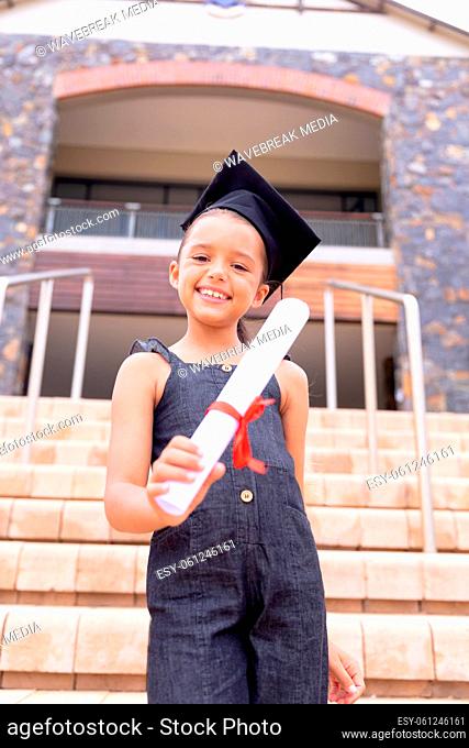 Portrait of smiling caucasian elementary schoolgirl with mortarboard showing degree certificate