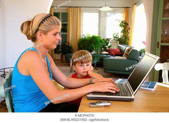 young mother with son, working at a laptop. The boy stands beside the table, looking bored and sad