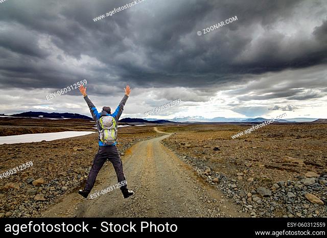 Happy Sucessful Winning Hiking young Man Jump with Backpack and raised arms on Trail road. Hiking in arctic desert of Iceland on Road F550