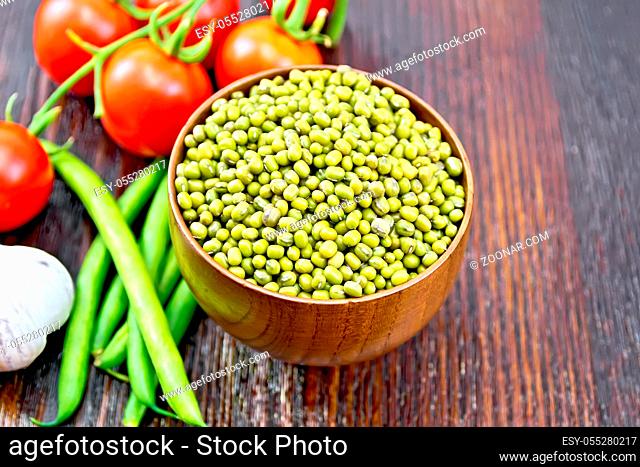 Green lentils mung in a bowl, pods of beans, garlic and red tomatoes on a wooden board background