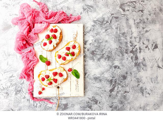 Three delicious toasts with Ricotta cheese and raspberries, decorated with mint leaves on white vintage cutting board on light background. Top view