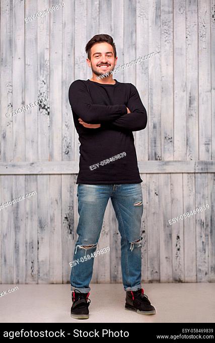 Brunette model man posing in full length with his arms crossed over white background. Man in black sweater and jeans smiling