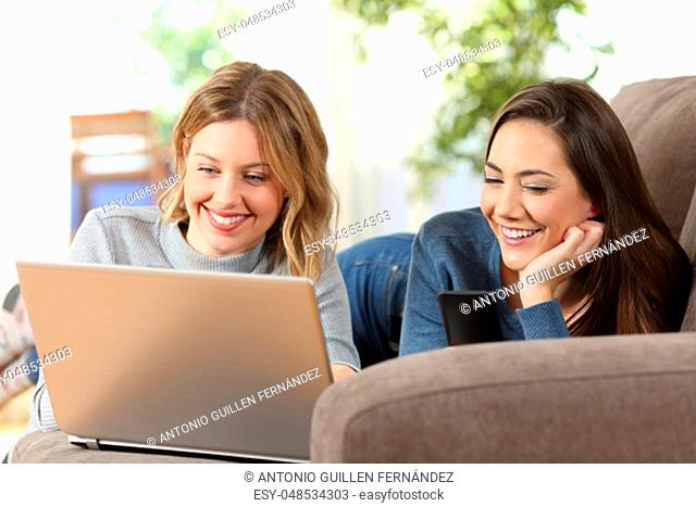 Front view portrait of two happy friends using a laptop and a smart phone lying on a couch at home