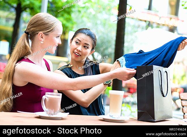 Pretty Asian woman showing a friend her purchase of a blue garment as they sit at a restaurant table enjoying refreshments
