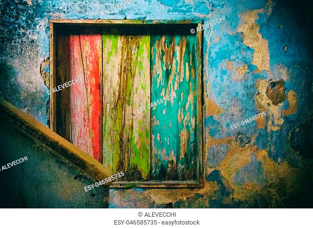 In a decayed ambient with cracked blue walls a wood internal window is painted in red, yellow and green in front of a staircase