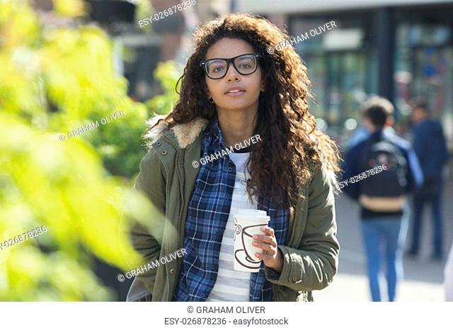 A happy female student looks at the camera as she walks through the city. She is carrying her hot drink and is wrapped up warm for the colder weather