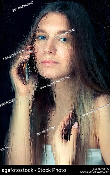 Portrait of young blonde woman with phone hands. Female behind the window glass with raindrops. Self-isolation lockdown concept