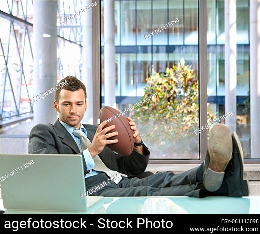 Relaxed businessman sitting at desk in front of office windows, holding football and smiling