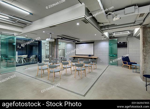 Zone for presentations in office in a loft style with brick walls and concrete columns. Zone has many gray chairs and a projector above them